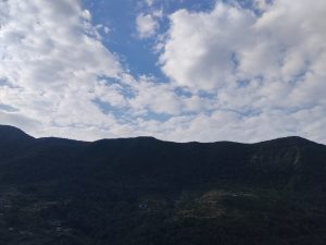 A scenic view of a mountain range under a sky filled with scattered white clouds. The mountains are covered in lush greenery, and there are small buildings visible at the foot of the hills.
