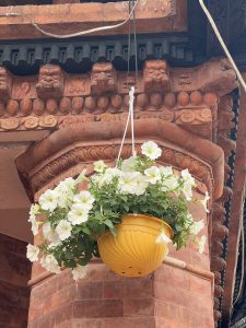 A yellow hanging flower pot with white petunias is suspended in front of an ornately carved brick pillar.