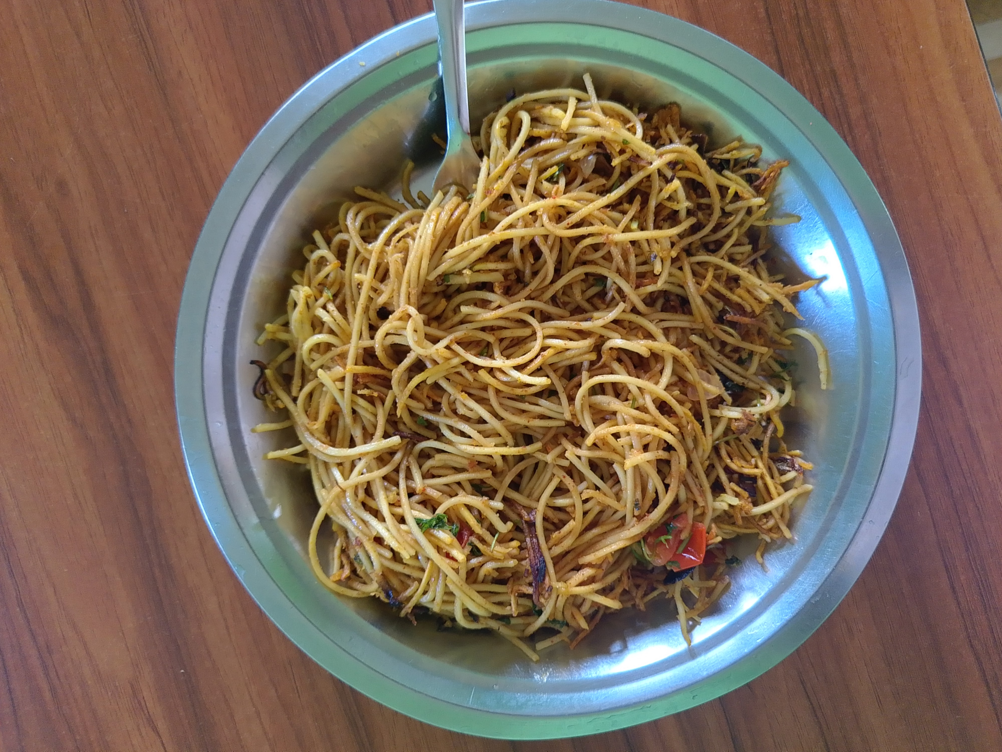 A plate of cooked chowmein with vegetables, including bell peppers and onions, served on a metal plate with a fork on a wooden table.
