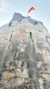 A close-up view of a stone tower, showcasing rough, uneven stonework in various shades of gray and brown. At the top of the tower, a bright orange flag flutters against a pale sky