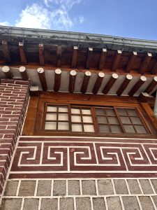 The image shows a portion of a building's exterior, featuring a wooden overhang supported by rounded beams. Below the overhang, there is a window with multiple rectangular panes, framed in wood.