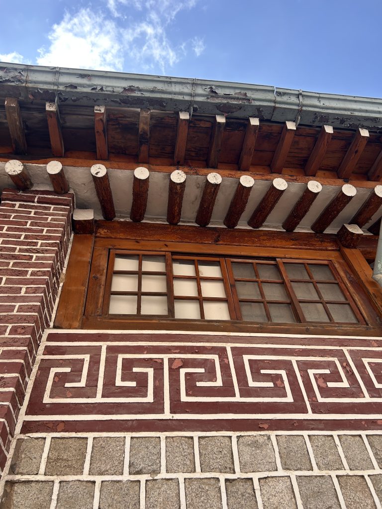 The image shows a portion of a building’s exterior, featuring a wooden overhang supported by rounded beams. Below the overhang, there is a window with multiple rectangular panes, framed in wood.
