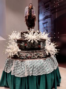 Statue adorned with a crown and wings stands atop an ornate pedestal surrounded by white floral arrangements. The pedestal is covered in decorative carvings and draped with lace and green fabric. #WCNP2025