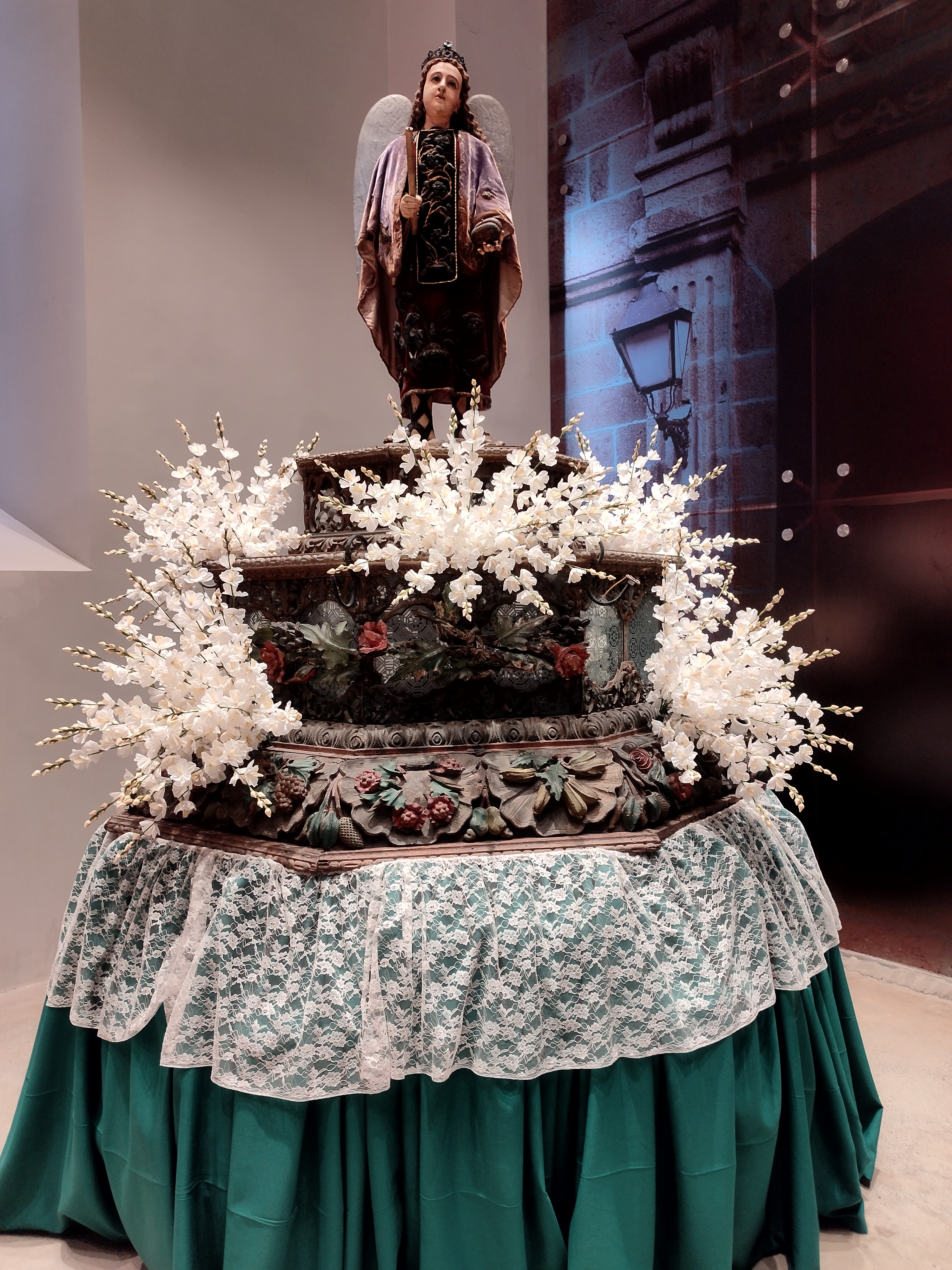 Statue adorned with a crown and wings stands atop an ornate pedestal surrounded by white floral arrangements. The pedestal is covered in decorative carvings and draped with lace and green fabric. #WCNP2025