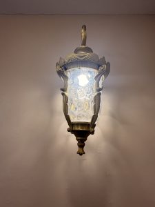 An ornate wall-mounted lantern with a decorative metal frame and textured glass, illuminated against a plain wall.