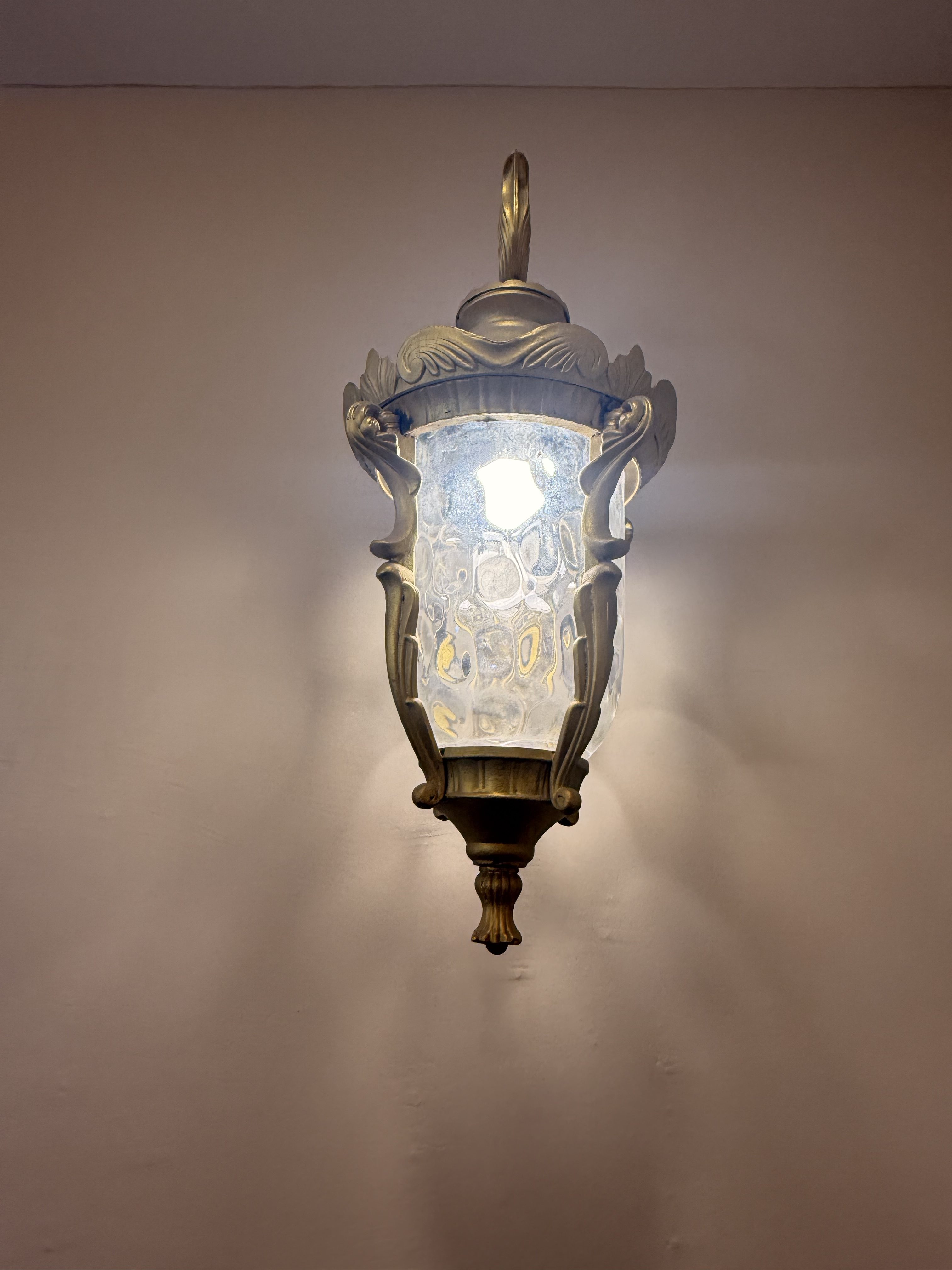 An ornate wall-mounted lantern with a decorative metal frame and textured glass, illuminated against a plain wall.