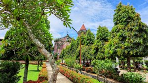 A lush garden scene with various green trees and shrubs lining a paved walkway. In the background, a historical church with a weathered facade and a red-tiled bell tower is partially visible. 