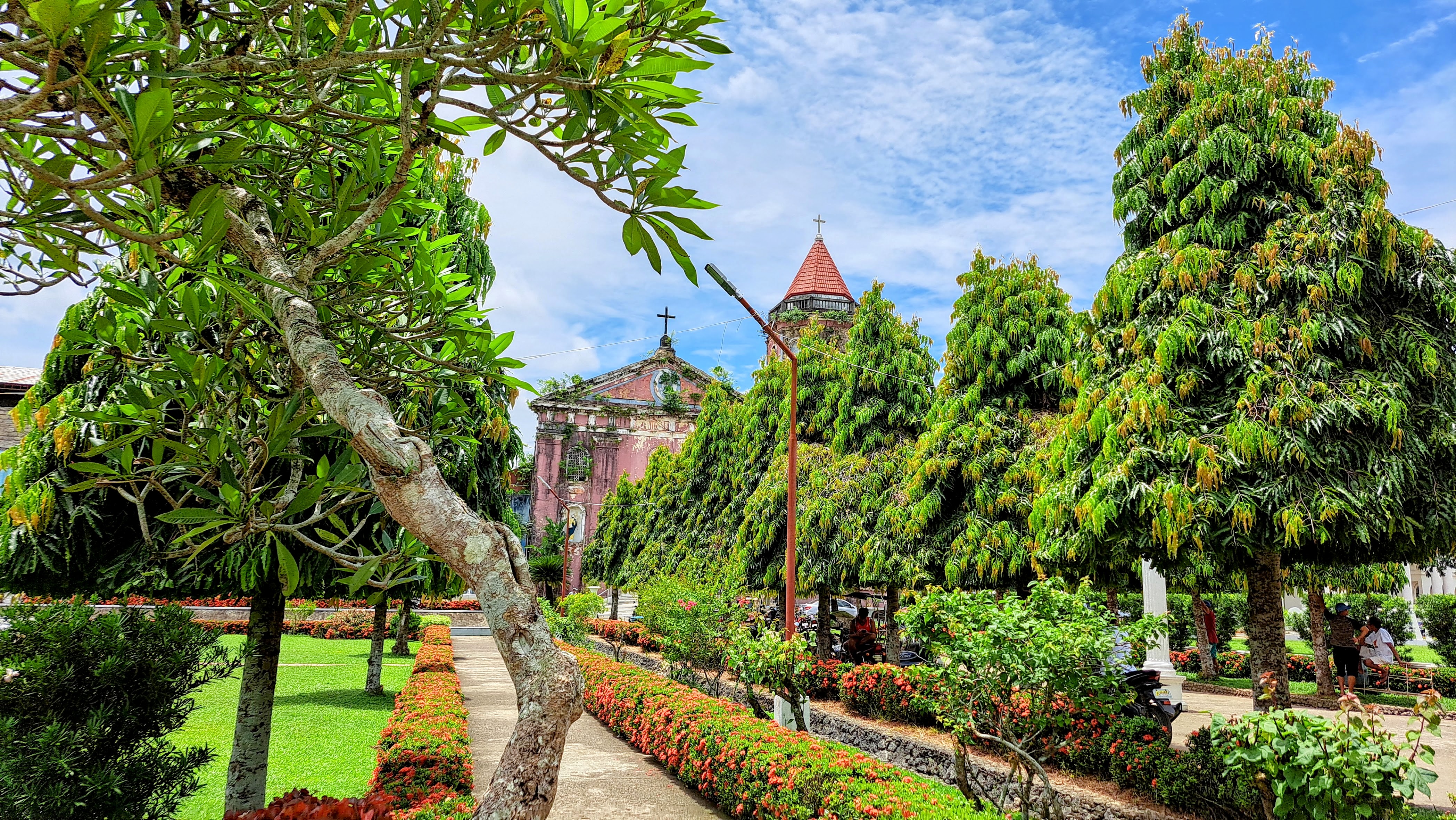 A lush garden scene with various green trees and shrubs lining a paved walkway. In the background, a historical church with a weathered facade and a red-tiled bell tower is partially visible.