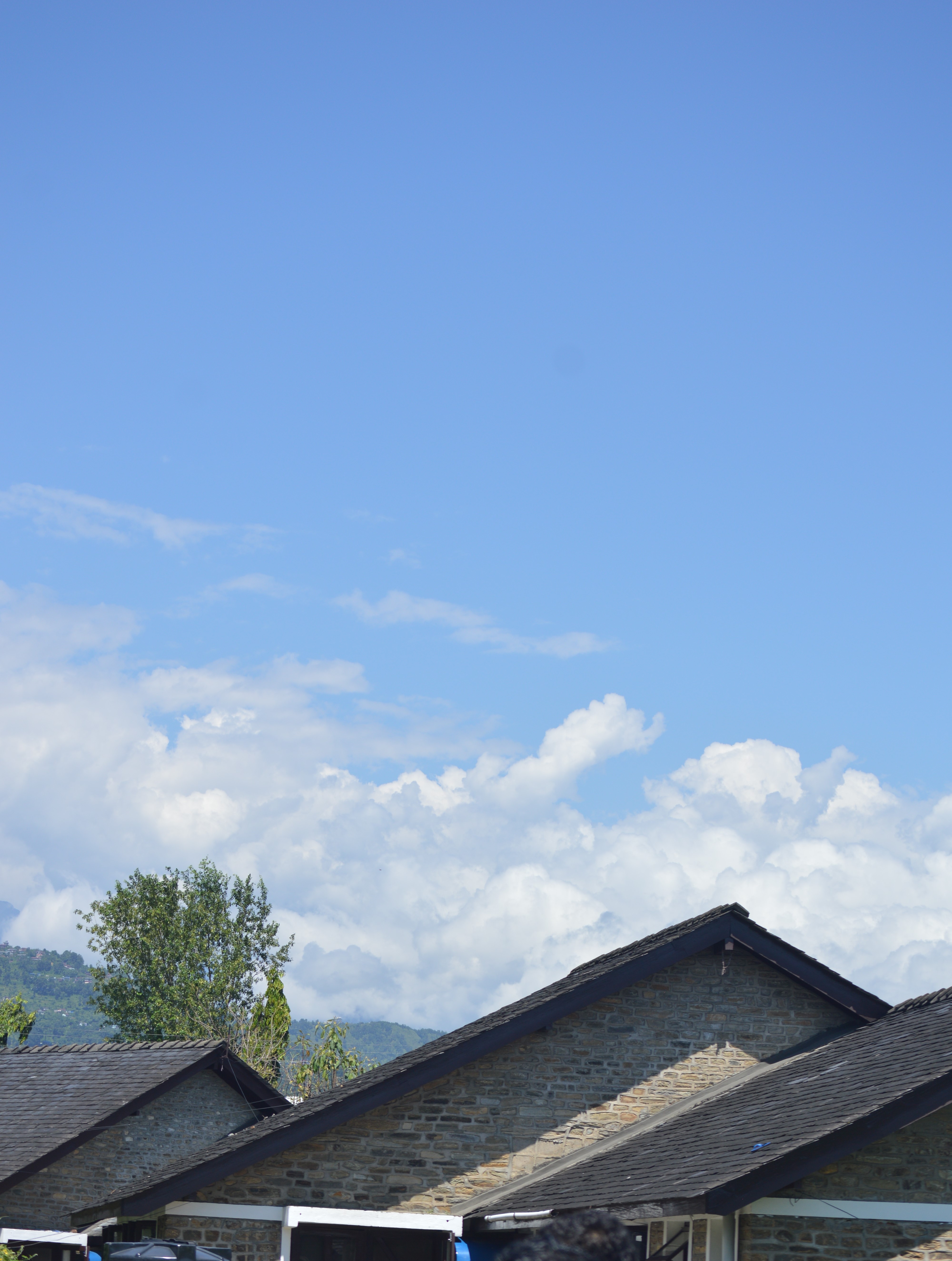 A row of stone houses with dark shingles and pitched roofs under a bright blue sky. In the background, fluffy white clouds and a green tree are visible.