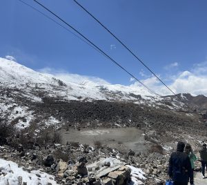 A scenic view of a mountainous landscape featuring snow-capped peaks under a clear blue sky. In the foreground, rocky terrain and patches of snow are visible, along with sparse vegetation and a small dry area.