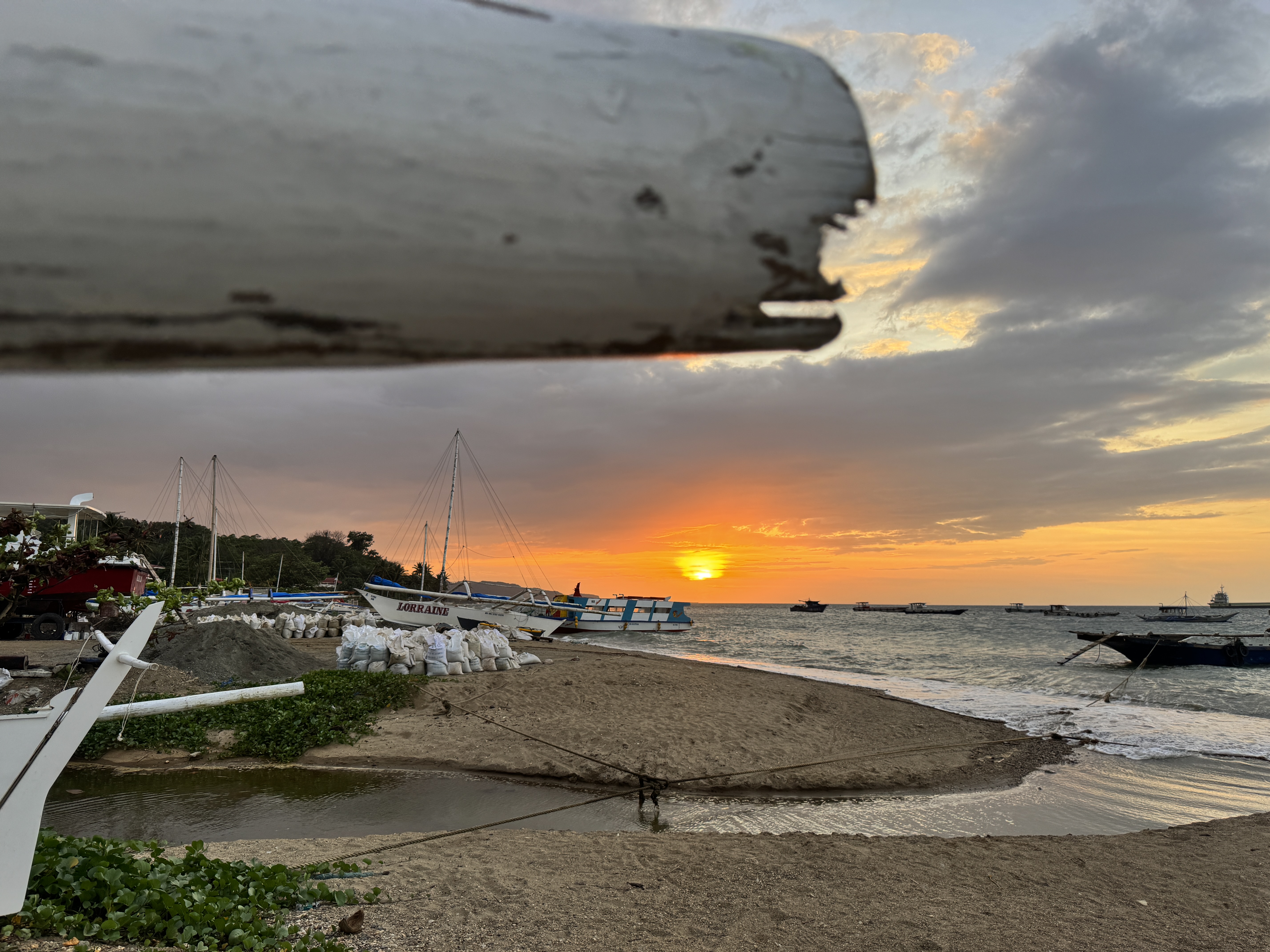 A beautiful sunset over the ocean with boats docked near a sandy shore, piles of white bags, and green vegetation in the foreground.