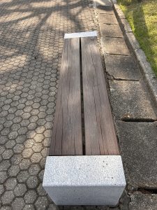 A simple wooden bench with two long, dark wooden planks as the seating surface, supported by light-colored stone end pieces.