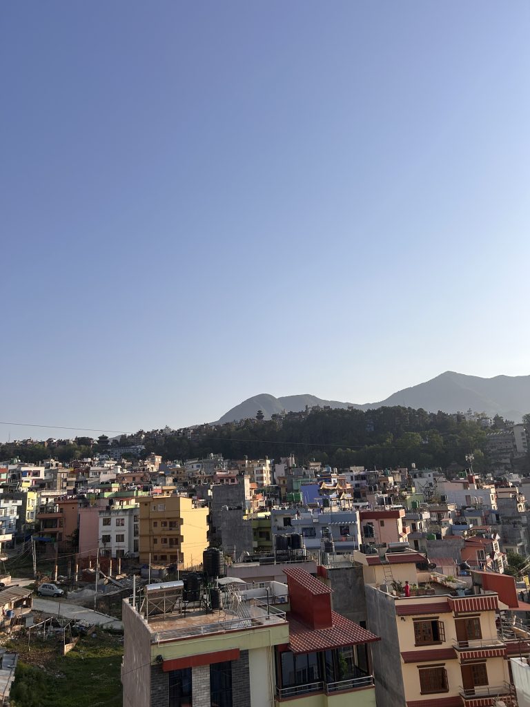 A cityscape view showing densely packed buildings with colorful roofs. In the background, there are forested hills and a clear blue sky.