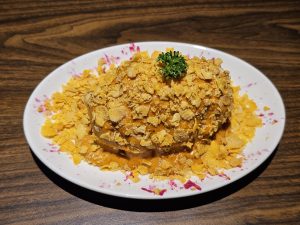 Hasselback Potato Chicken coated in crushed cornflakes and topped with a sprig of parsley, served on a white plate with a few red specks. The plate sits on a wooden surface. Captured from Kozhikode, Kerala.