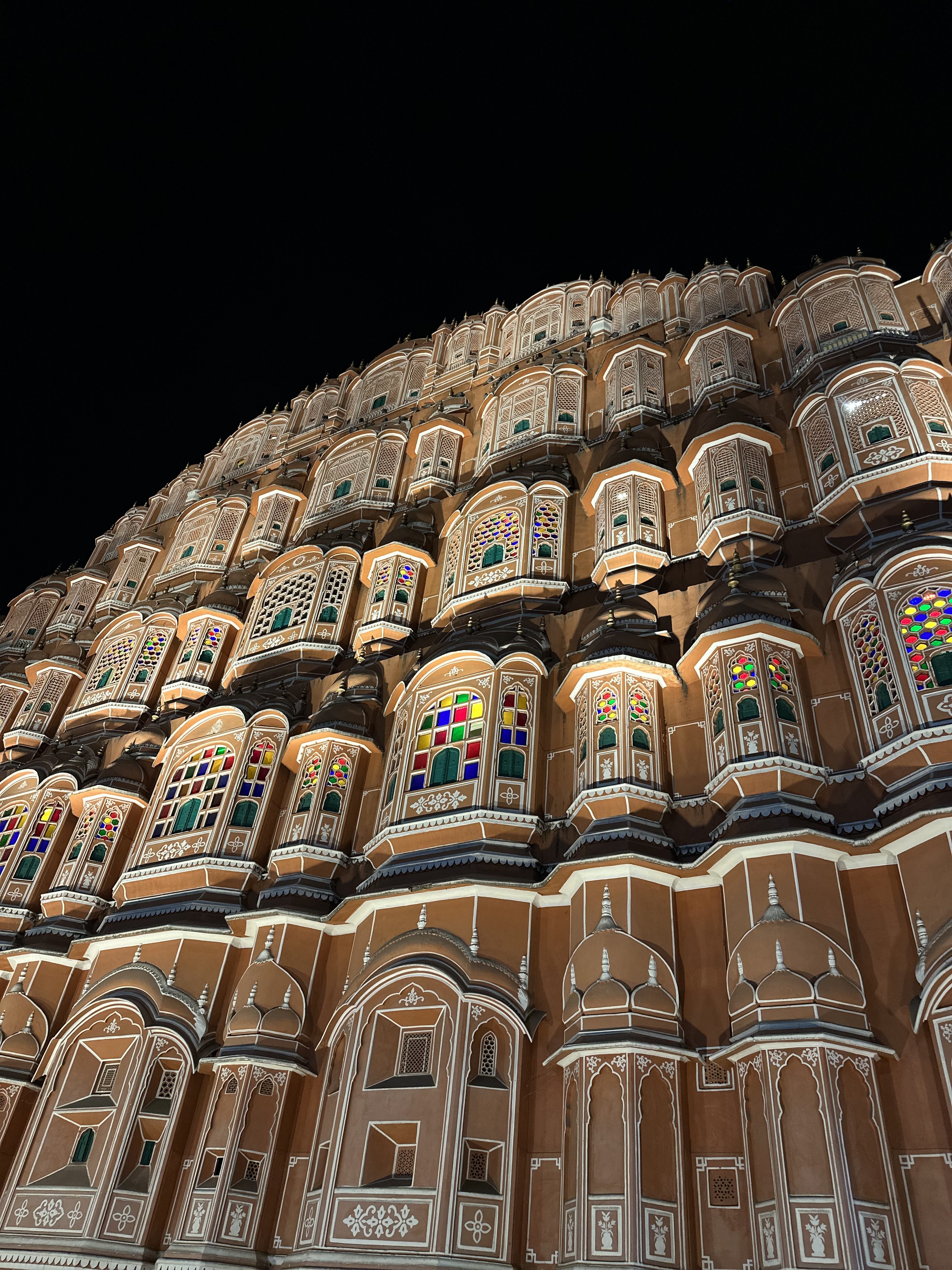 An ornate building facade with multiple small windows featuring colorful stained glass and intricate architectural details, illuminated against a dark night sky.