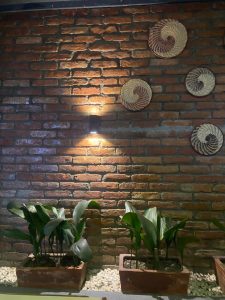 A brick wall decorated with spiral-patterned woven baskets. Below, two rectangular planters contain tall green plants, set on a bed of white pebbles. A small wall light casts a warm glow on the bricks.