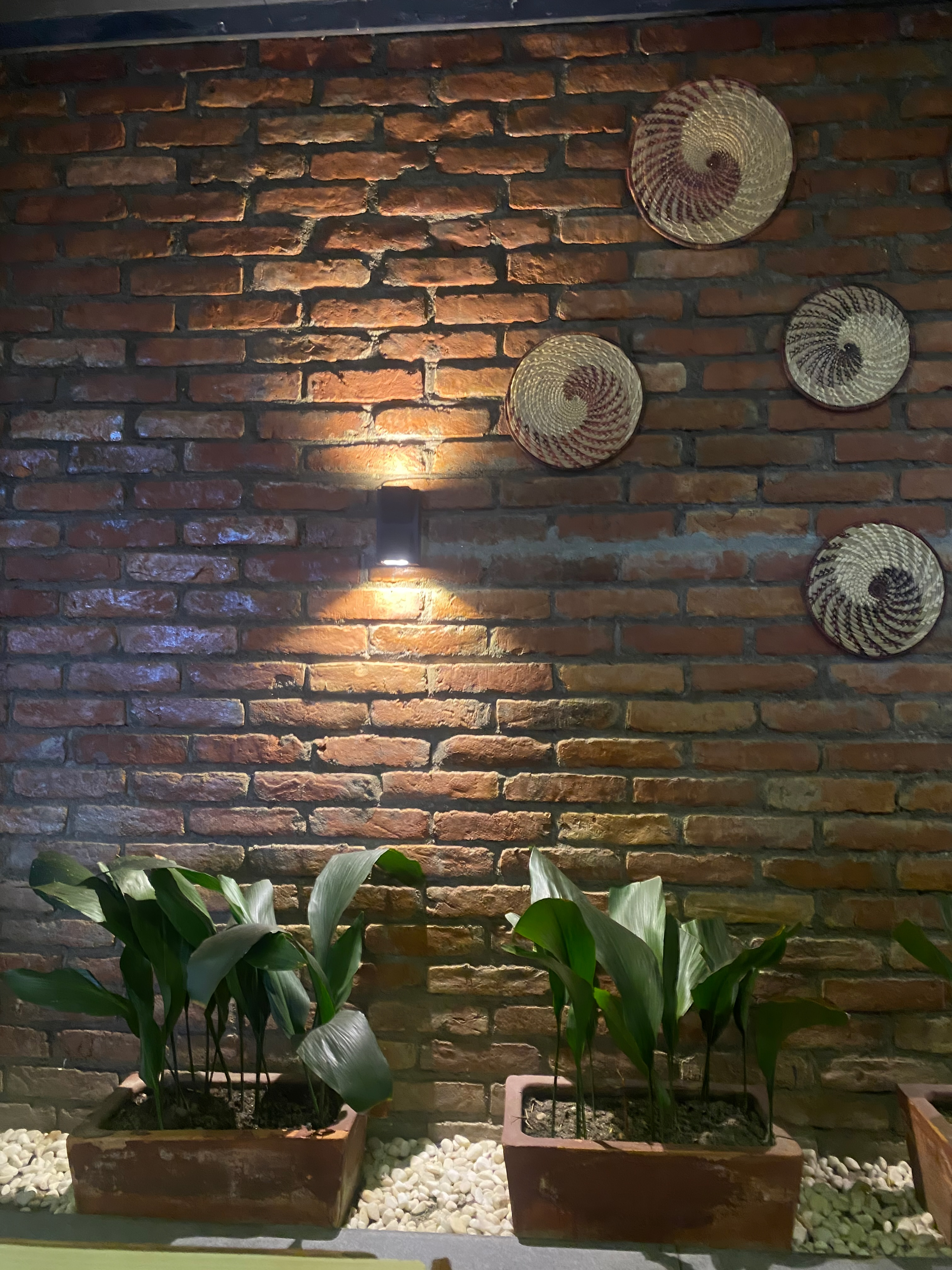 A brick wall decorated with spiral-patterned woven baskets. Below, two rectangular planters contain tall green plants, set on a bed of white pebbles. A small wall light casts a warm glow on the bricks.