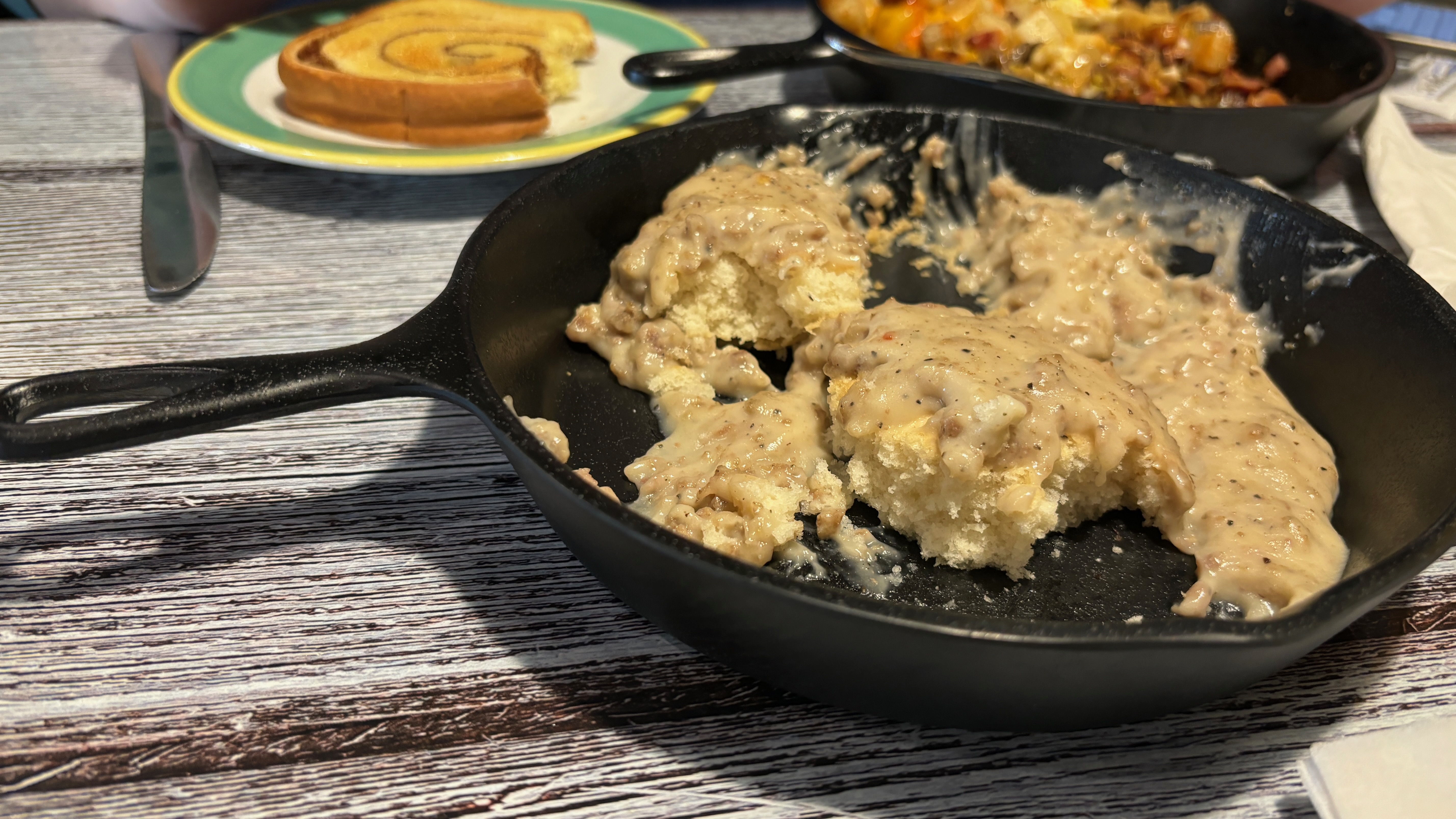 Closeup of American biscuits and gravy in a cast iron pan. Cinnamon swirl toast and breakfast casserole in the background.