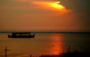 A silhouetted boat floats on a calm body of water during sunset, with the sky and water glowing in shades of orange and red under a partially cloud-covered sun.