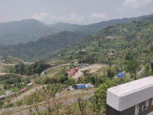 A scenic view of a mountainous landscape with lush green hills and scattered buildings, as seen from a roadside. A winding road leads through the valley, and the sky is partially cloudy.