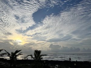 Scenic view of a beach at sunset with dramatic cloud patterns in the sky, palm trees silhouetted in the foreground, and a few people standing near the shoreline.