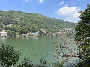 A scenic view of Lake, surrounded by lush green hills. The lake is calm and dotted with small boats, and colorful buildings line the hillside across the water. A partly cloudy blue sky adds to the serene atmosphere, with trees framing the foreground.

