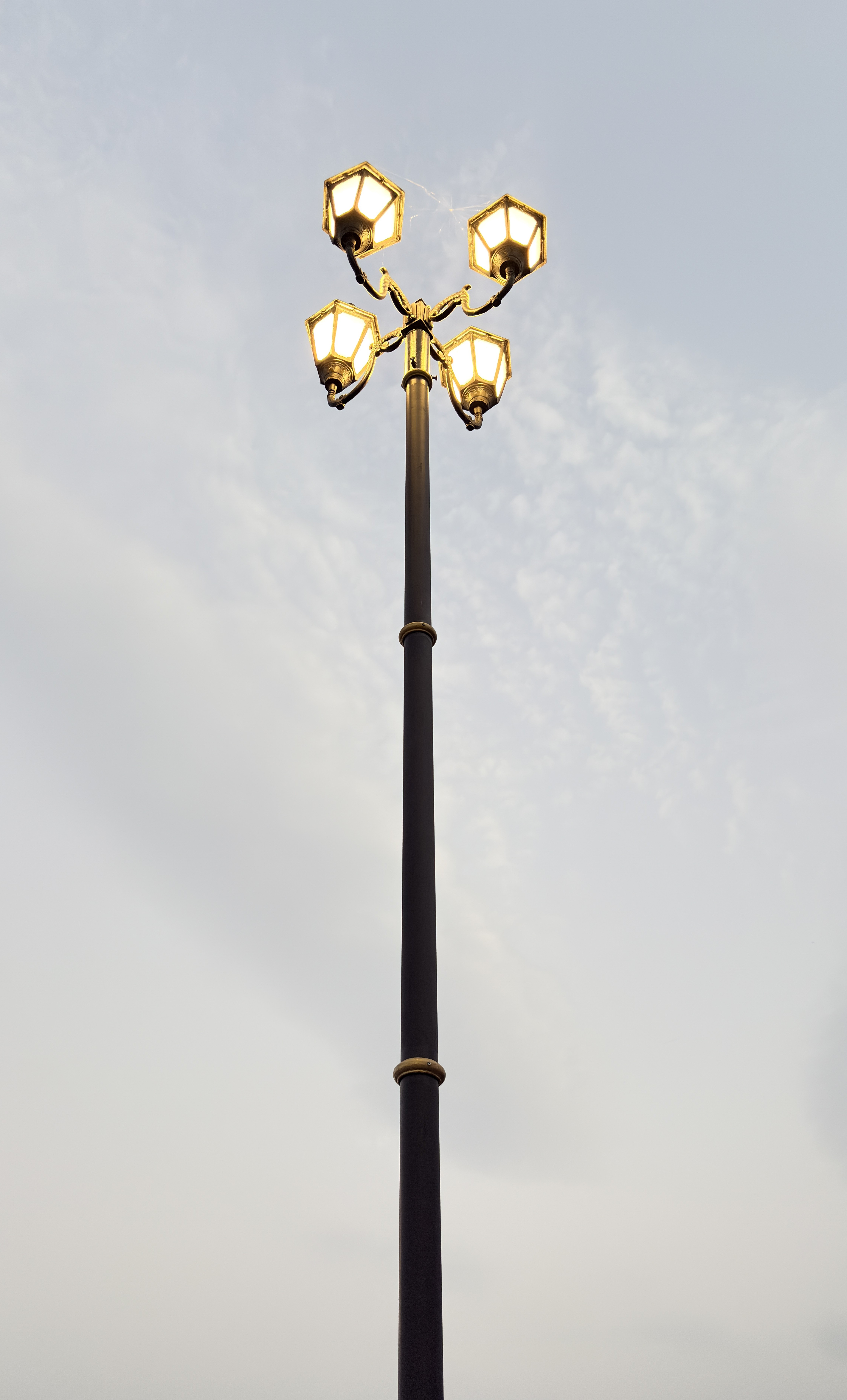 A tall street lamp with four illuminated lanterns arranged in a circular pattern against a cloudy sky.