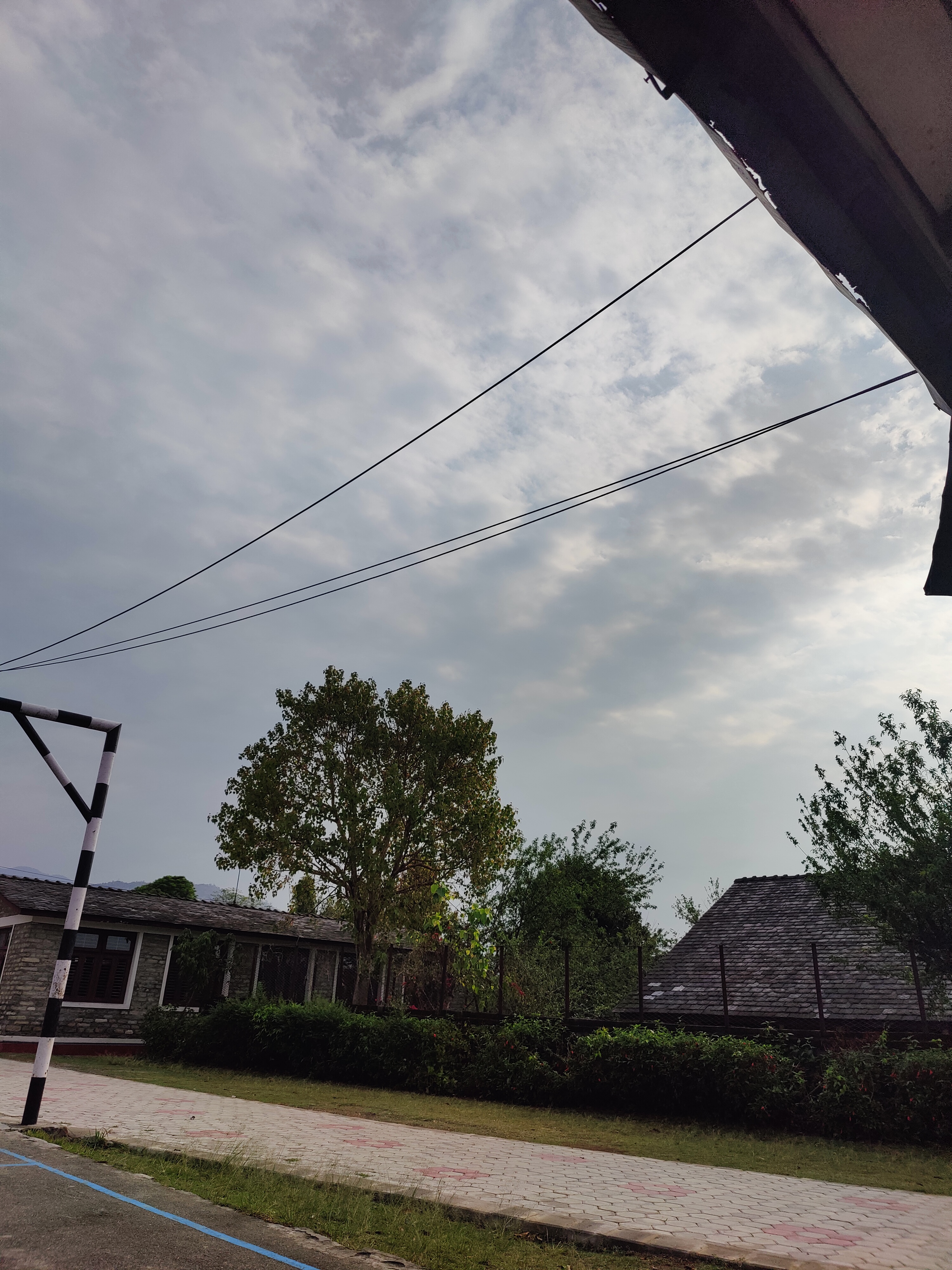 A cloudy sky with overhead power lines above a quiet area featuring trees, a paved walkway, and buildings with sloped roofs surrounded by greenery.