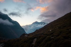 A scenic mountain landscape at dusk with clouds draped over the valley and peaks in the distance. The sky features a gradient of blue and orange hues, while the foreground displays a sloped, grassy terrain.