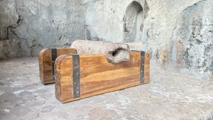 A wooden crate-like structure sits on a textured stone floor inside a stone-walled space. The crate has a rounded top and is reinforced with metal straps. Inside the crate, there is a cylindrical stone artifact that appears weathered, suggesting it may be an old cannon or similar object
