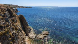 A scenic coastal view featuring rugged cliffs on the left side, with sparse vegetation. The ocean is calm with sparkling sunlight reflecting off the water. In the distance, land extends into the sea, with a few buildings visible.
