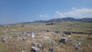 A vast landscape featuring Pamukkale ancient city ruins scattered across grassy terrain, with remnants of stone structures and columns in the foreground. In the background, hills rise under a clear blue sky, with a few clouds visible. The scene conveys a sense of history and natural beauty in an archaeological site.

