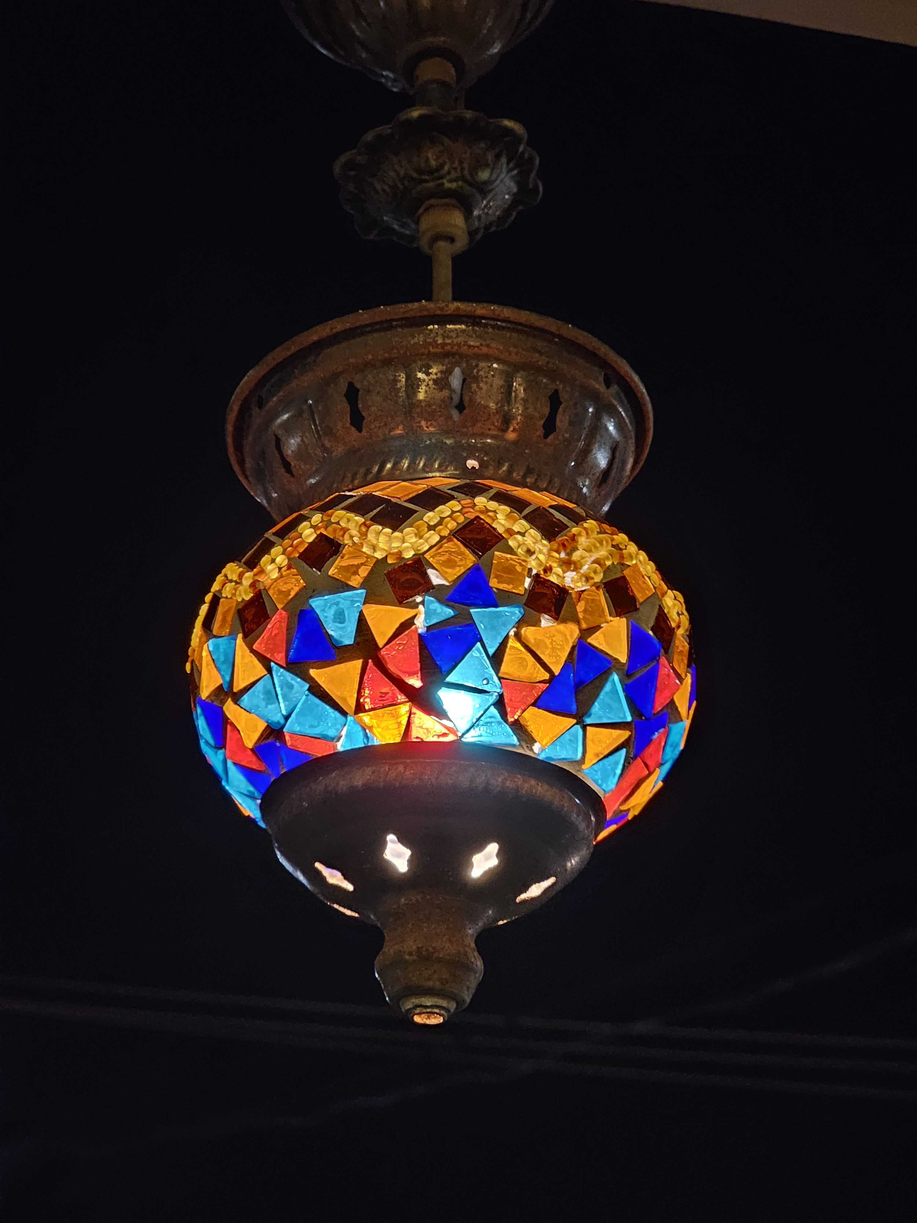 A Turkish mosaic lamp hanging from a ceiling fixture against a dark background. The lamp features a spherical lampshade adorned with colorful, geometrically shaped glass pieces, and it emits a warm glow, highlighting its intricate design and handcrafted details. Captured from Kozhikode, Kerala.