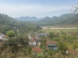 A scenic view of a rural landscape with a cluster of houses scattered among lush greenery. Rolling hills covered with dense forests rise in the background under a clear blue sky