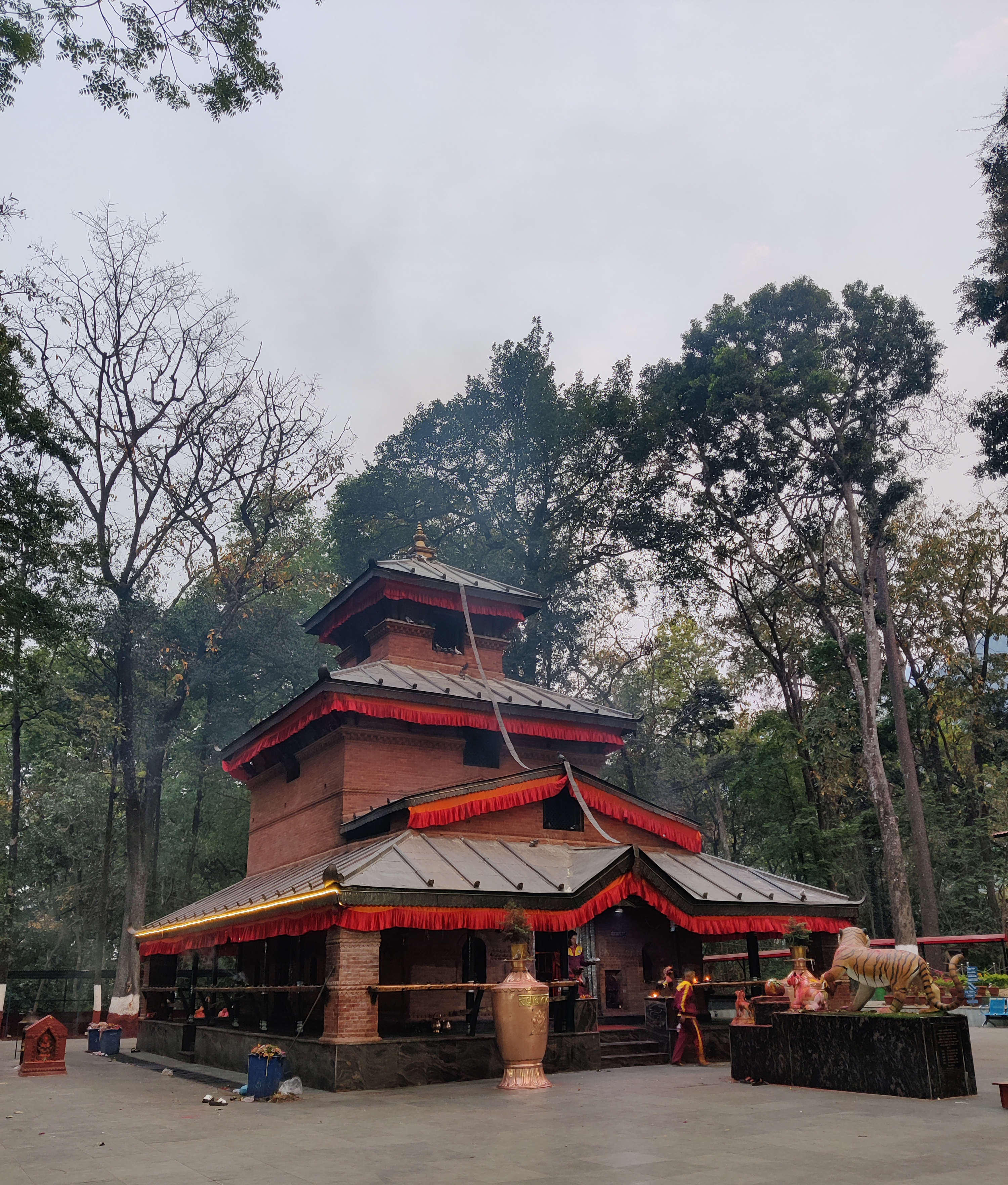A traditional multi-tiered temple with red trim details stands amidst tall trees. In front of the temple, there are large vases and a lion statue on a pedestal. The area is paved, and a few people are seen near the entrance, possibly participating in a ritual.