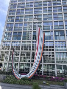 A colorful, curved sculpture with stripes in red, blue, orange, and white is positioned in front of a modern glass building.