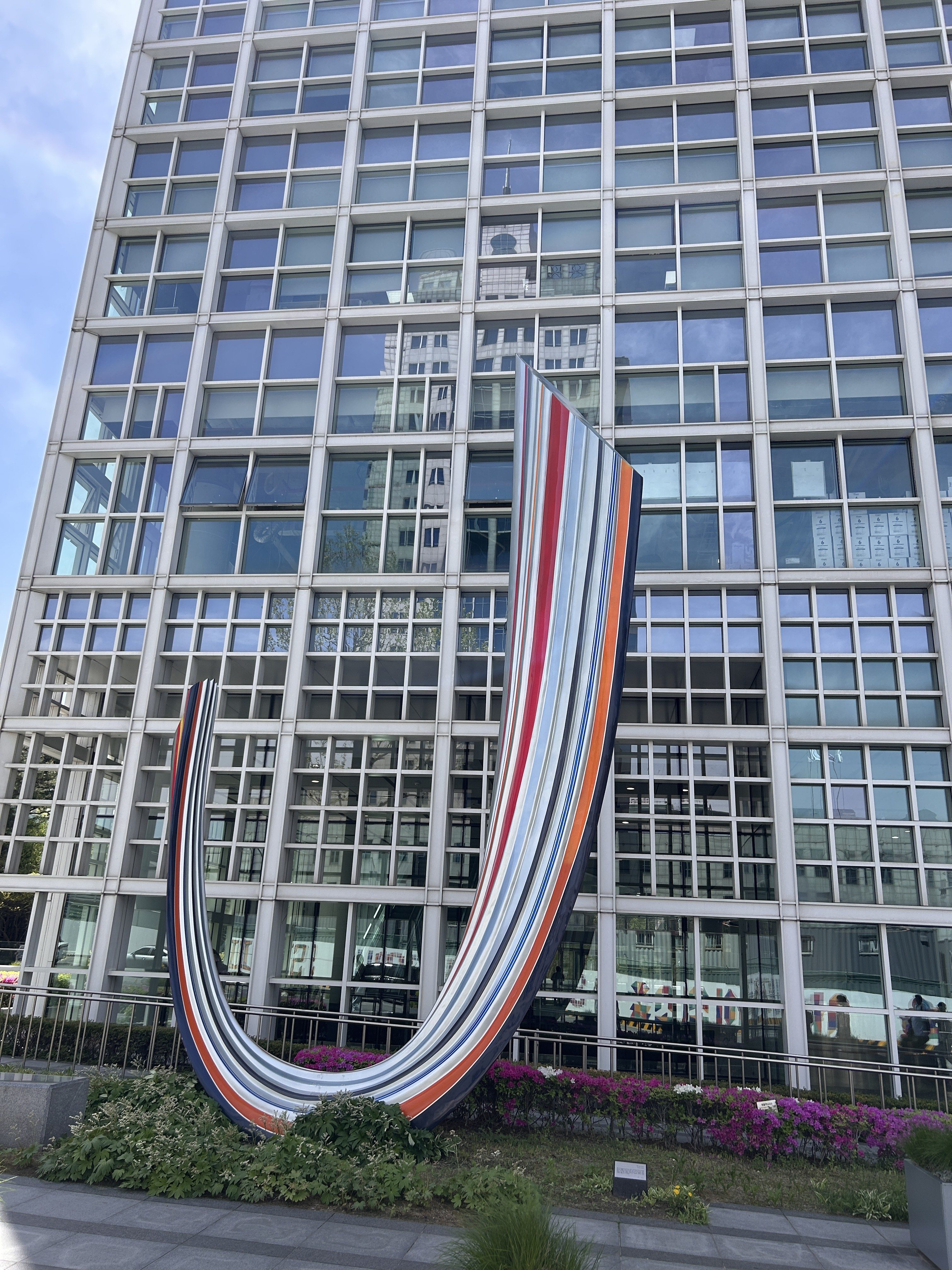 A colorful, curved sculpture with stripes in red, blue, orange, and white is positioned in front of a modern glass building.
