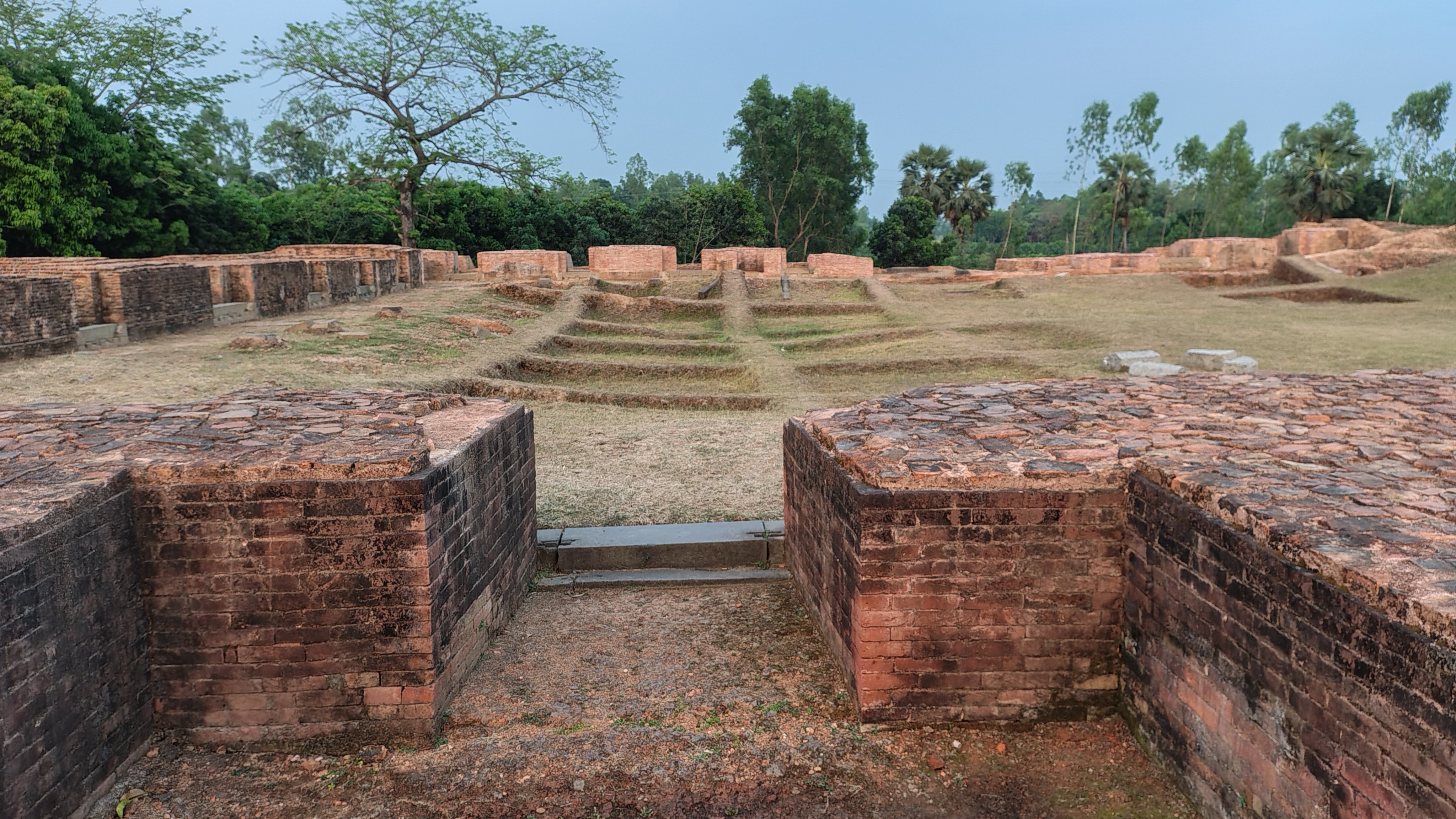 Ancient brick ruins set in a grassy area, surrounded by lush green trees. The structures are weathered and partially buried, with clear lines indicating former building foundations.