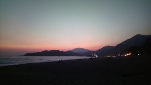 A serene beach scene at dusk, with soft pastel hues of pink and purple in the sky as the sun sets behind mountains.