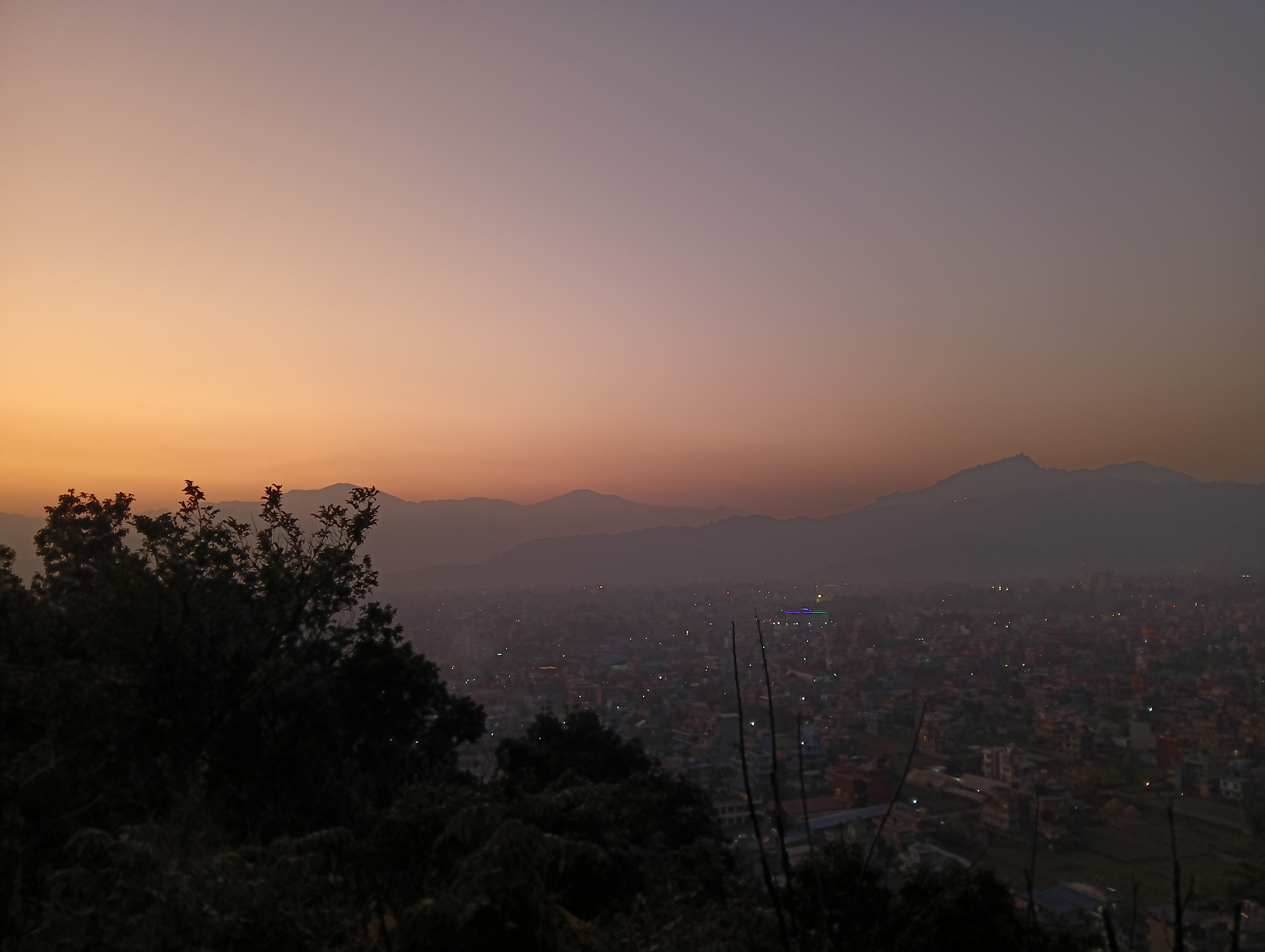 A cityscape at dusk with a hazy sky transitioning from orange to blue, silhouetted trees in the foreground, and mountains in the background.