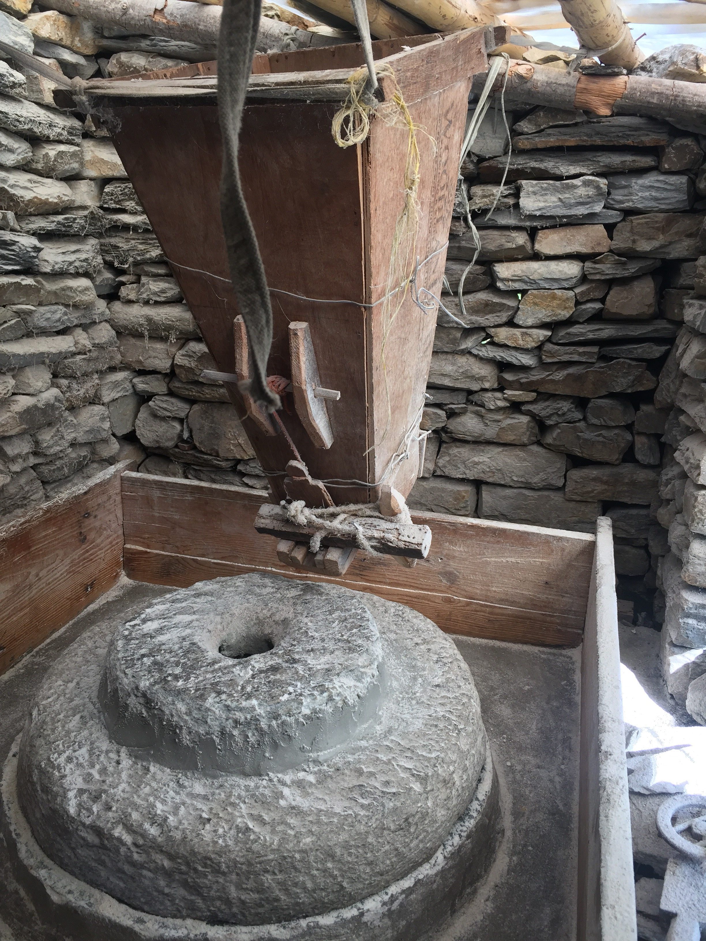 A traditional stone mill with a large circular grinding stone and wooden hopper, set within a rustic structure made of stone walls and wooden beams.
