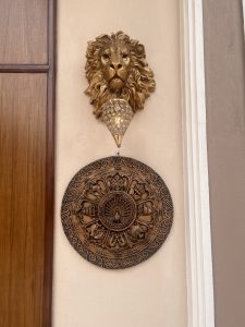 A decorative wall arrangement featuring a golden lion head sculpture at the top, a crystal-like hanging lamp below it, and an intricately carved wooden circular plaque with traditional motifs at the bottom. The setup is mounted on a beige wall next to a wooden door.