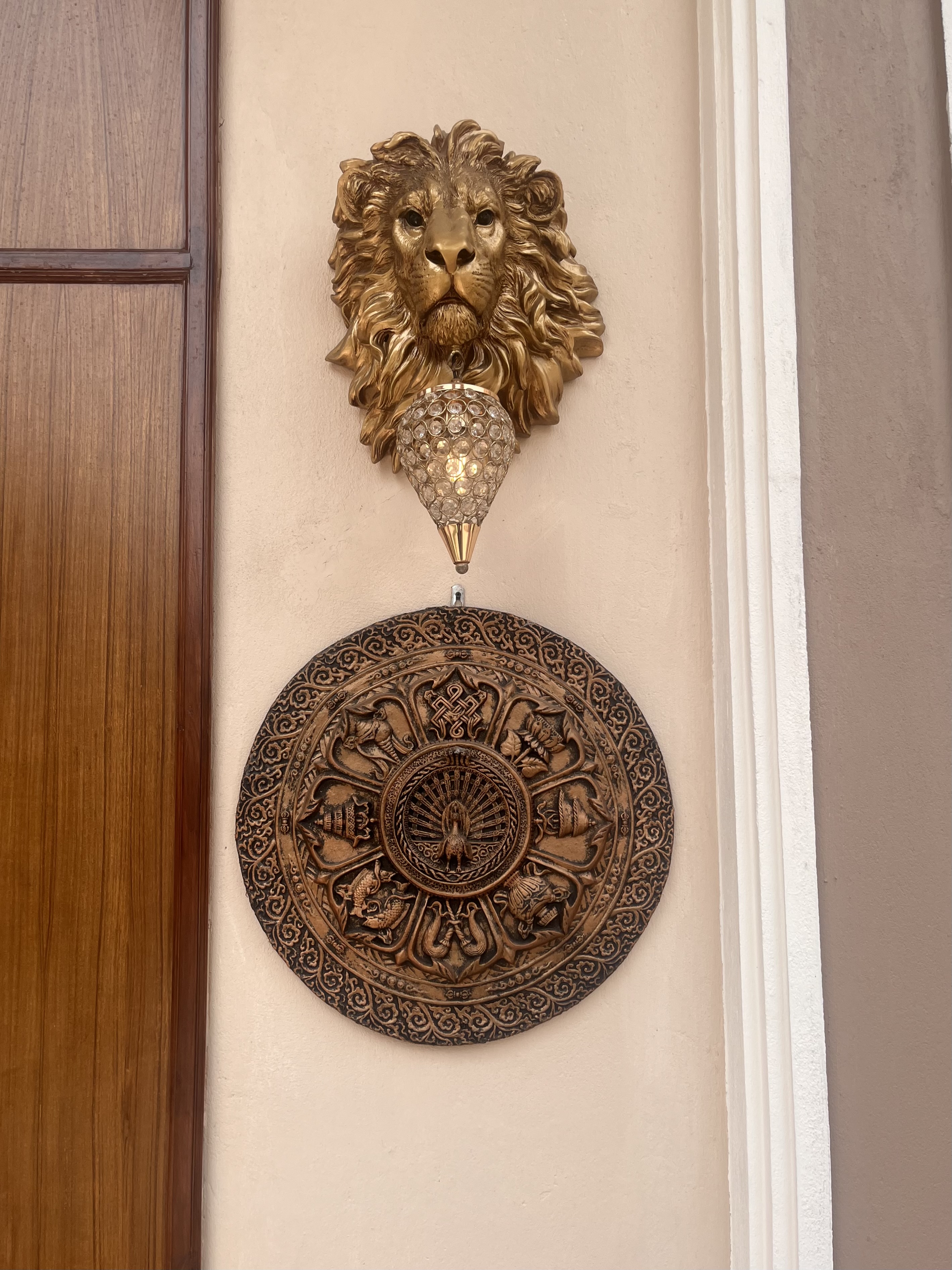 A decorative wall arrangement featuring a golden lion head sculpture at the top, a crystal-like hanging lamp below it, and an intricately carved wooden circular plaque with traditional motifs at the bottom. The setup is mounted on a beige wall next to a wooden door.
