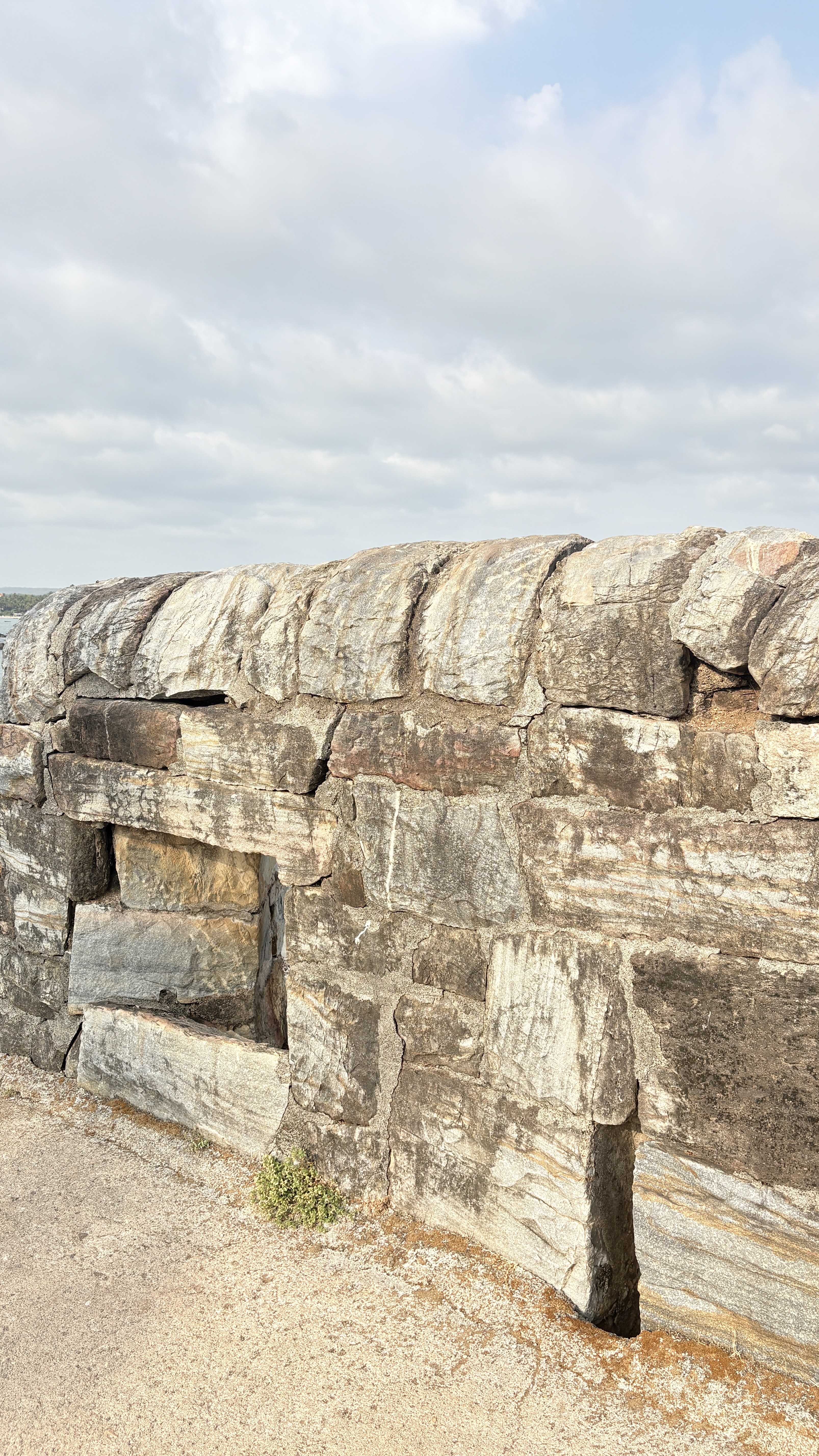 A weathered stone wall with large, uneven blocks and a small rectangular opening, set against a partly cloudy sky. Sparse green plants grow at the base of the wall, and the ground is covered with sandy soil.