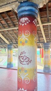 A brightly colored pillar under a wooden roof, painted with sections of red, yellow, and white. The pillar features intricate designs, including a prominent "Om" symbol in the center, surrounded by floral and swirl patterns. Other pillars in the background are similarly decorated, with a mix of colors and motifs, creating a vibrant and artistic at