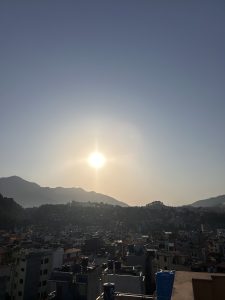 A scenic view of a city at sunset, with the sun setting over distant hills. The foreground features a cluster of buildings with varied rooftops, and the silhouette of the mountains is visible against a clear sky.
