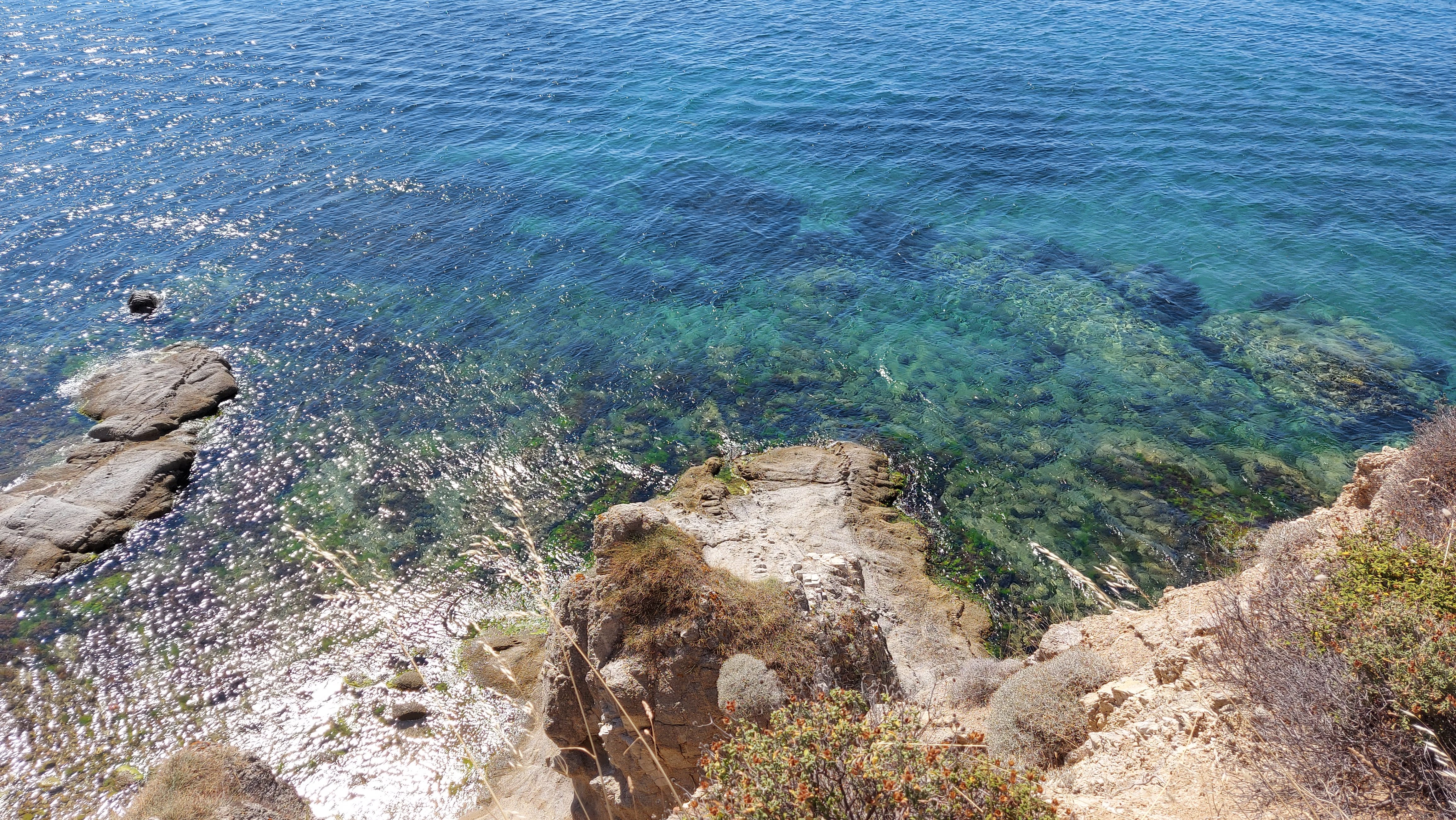 A coastline with clear blue water, rocky outcrops, and dry vegetation on the cliffside. Sunlight reflects off the water, illuminating the scene.