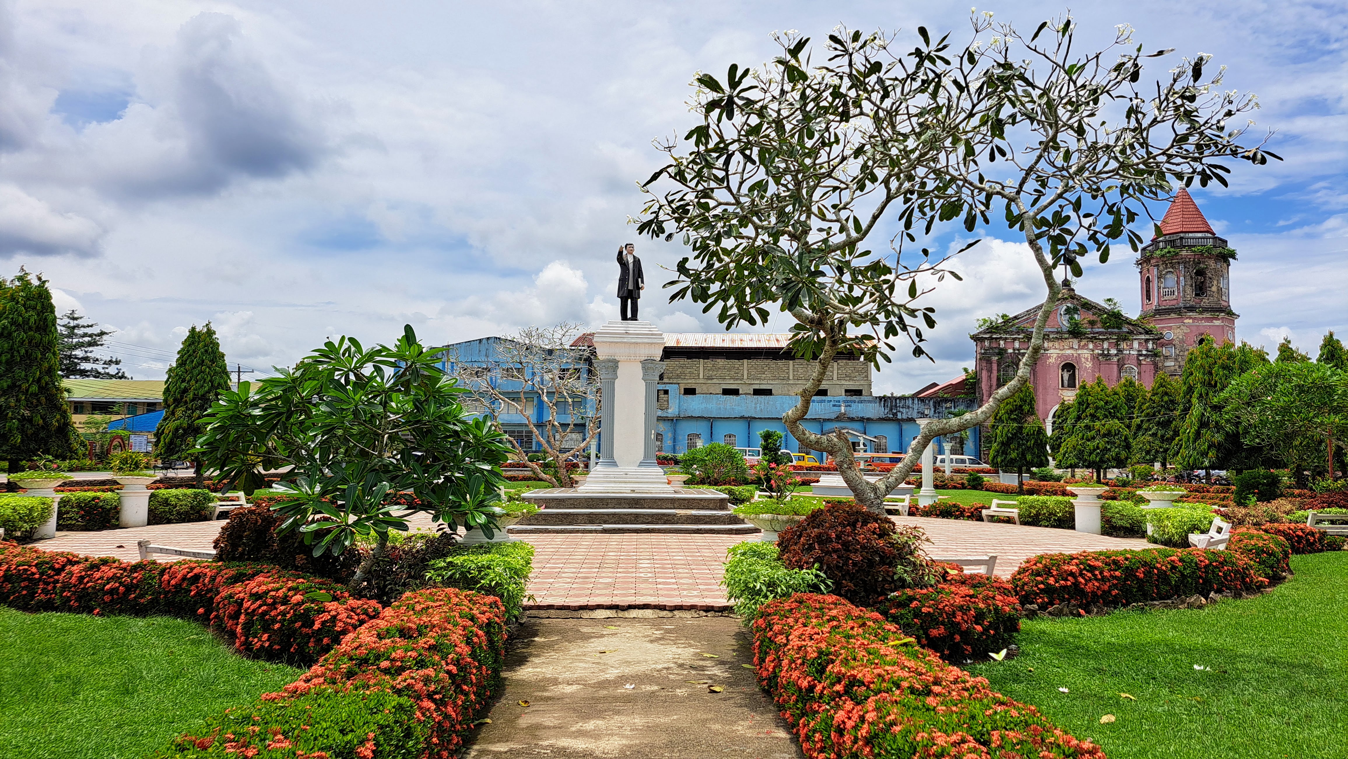 Aligned in quiet reverence, the path leads straight to the bust of José Rizal, standing watchful and proud, while the pink church on the right gently colors the backdrop with a sense of peace and history.