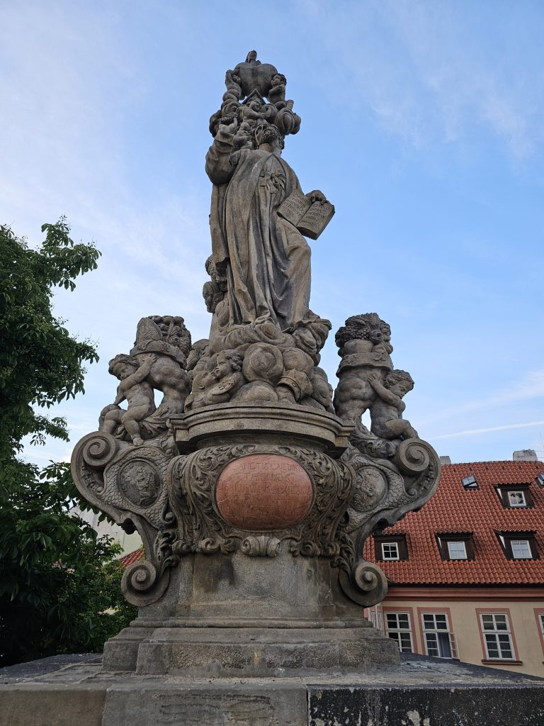 The statue of Saint Cajetan, located on the Charles Bridge in Prague. It is a baroque sculpture depicting the saint holding a book, surrounded by cherubs and ornate decorations, set against a backdrop of a clear sky and buildings with red-tiled roofs.