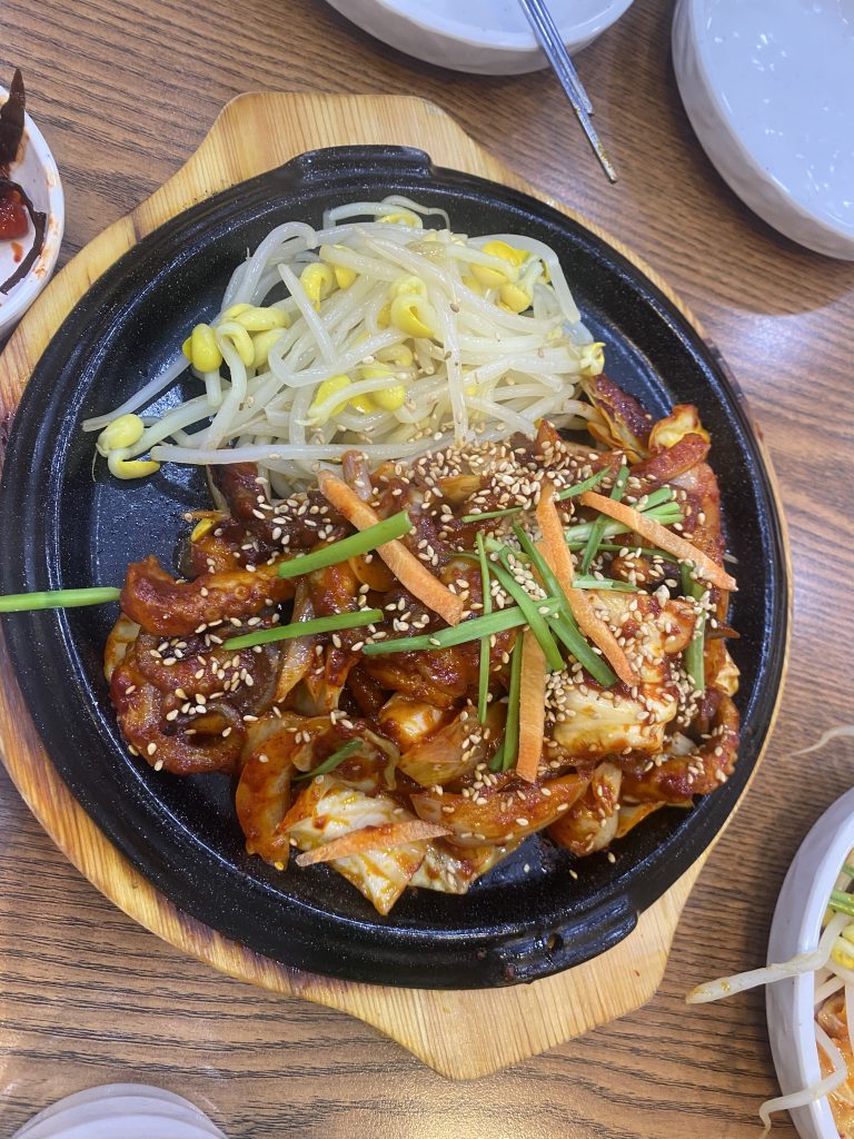 A sizzling hot plate of stir-fried food, featuring pieces of meat in a reddish-brown sauce, garnished with sesame seeds, green onions, and strips of carrot. Accompanying the main dish are bean sprouts and yellow mung beans.